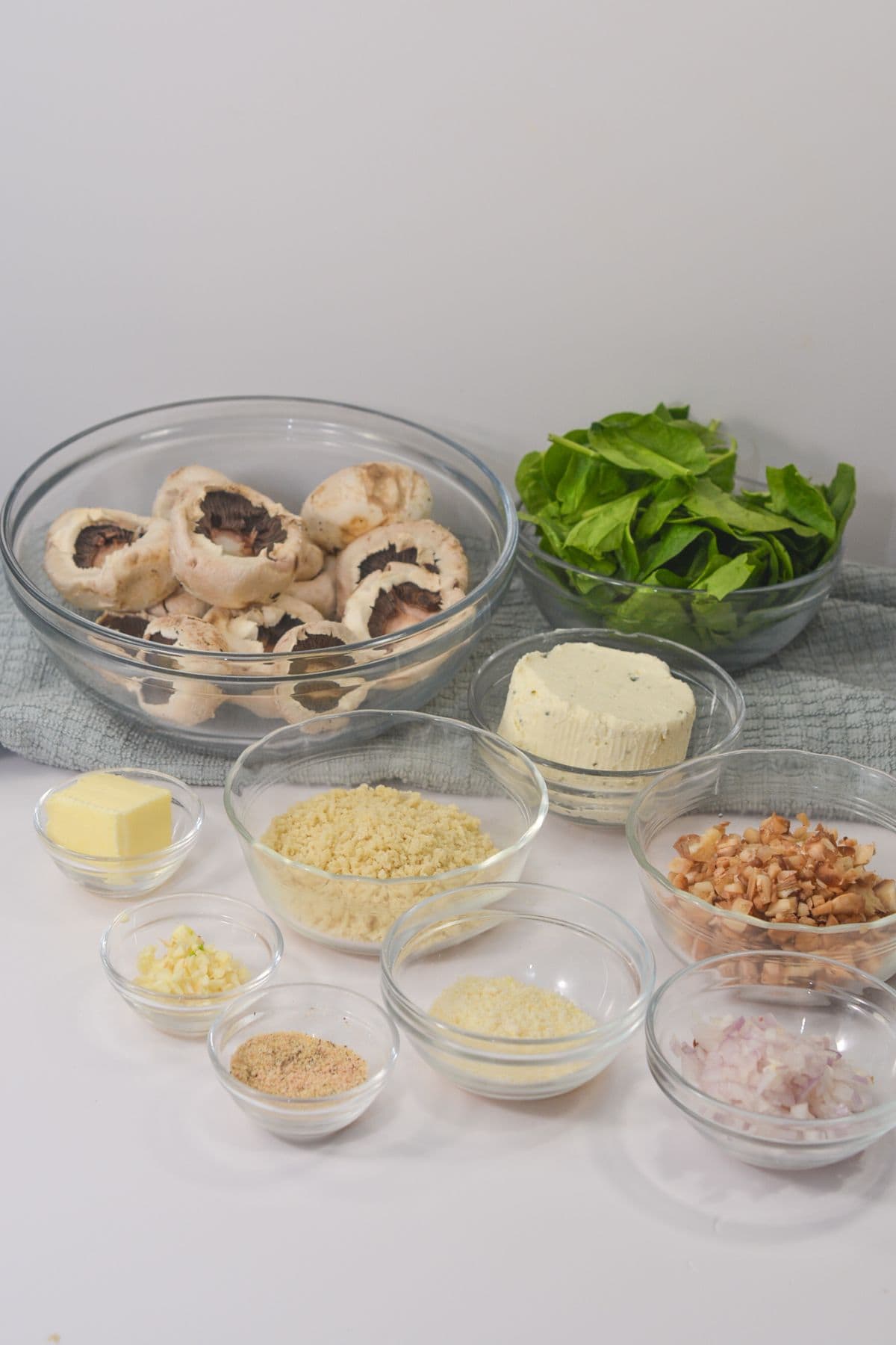 Various clear bowls containing butter, breadcrumbs, grated cheese, minced garlic, chopped nuts, and chopped onions arranged on a white surface.