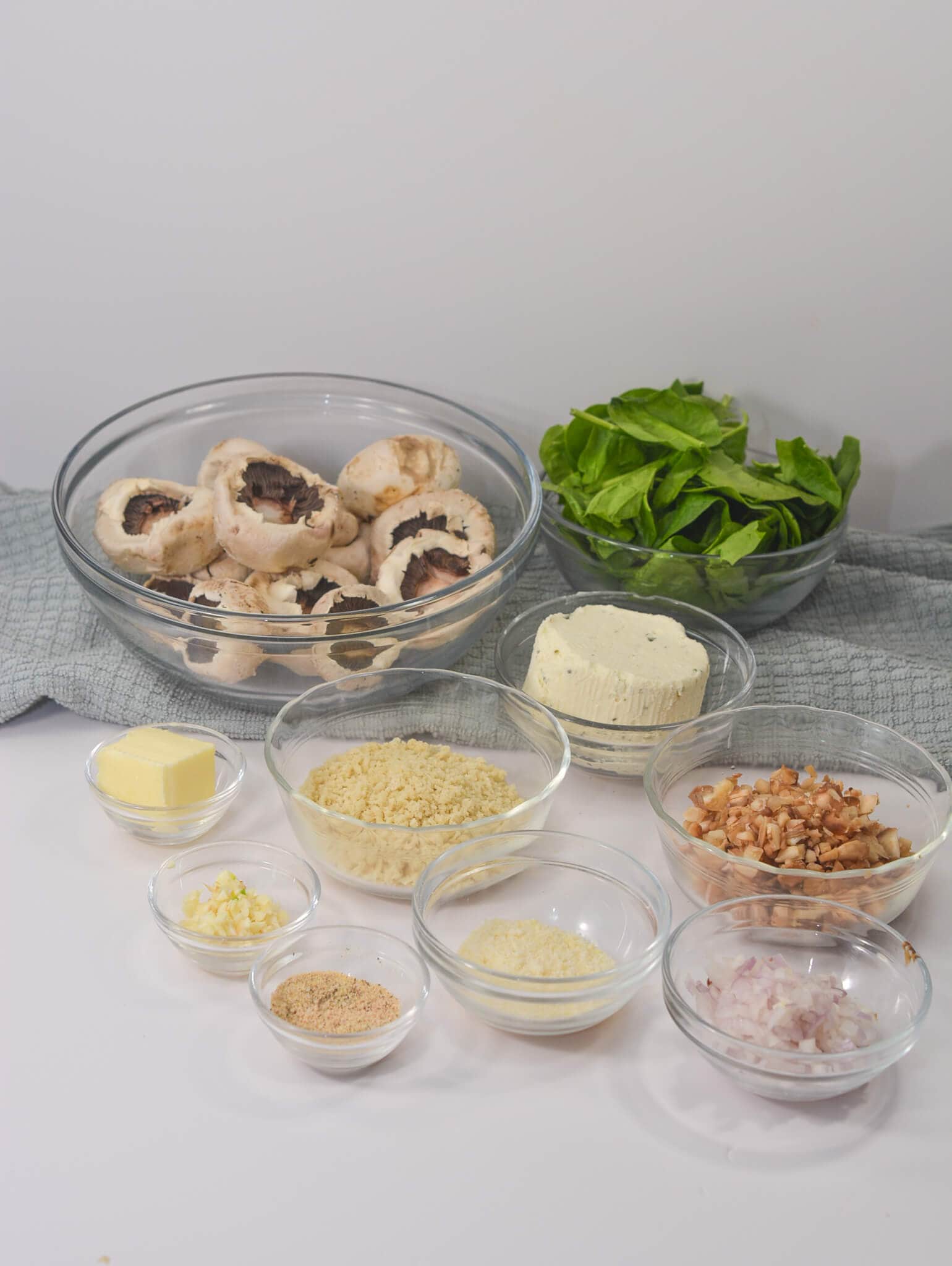 Ingredients including mushrooms, spinach, Boursin cheese, butter, and chopped nuts are perfectly arranged in bowls on a countertop.
