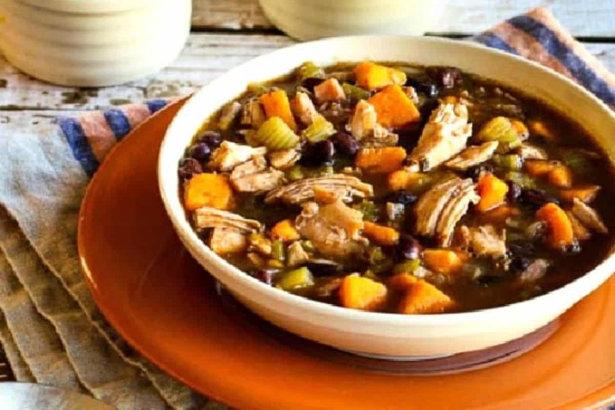 A close-up of a bowl of turkey, sweet potato, and black bean soup, served in a white bowl on a red plate.
