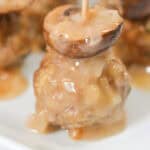 A veal marsala meatball topped with a mushroom cap, held together with a toothpick, is drizzled with savory sauce and artfully presented on a white plate.
