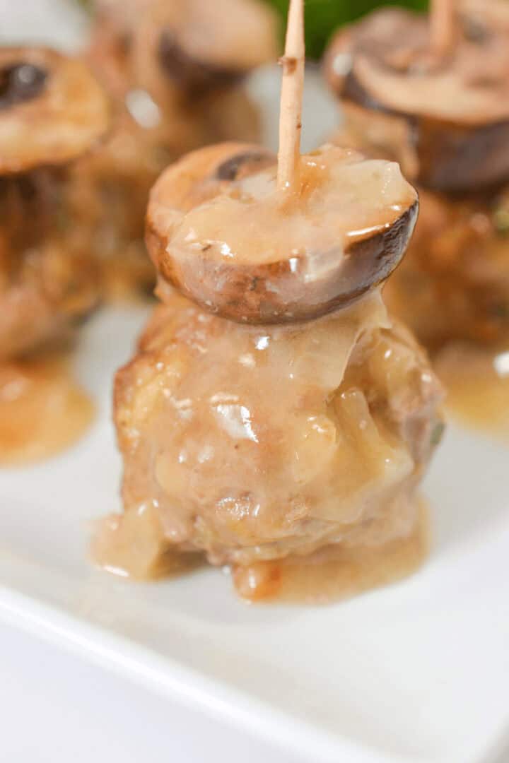 A veal marsala meatball topped with a mushroom cap, held together with a toothpick, is drizzled with savory sauce and artfully presented on a white plate.