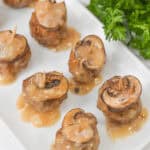 Savoury food with mushroom slices and skewered with toothpicks, drizzled with sauce, are arranged on a rectangular white plate. Fresh parsley is in the background.