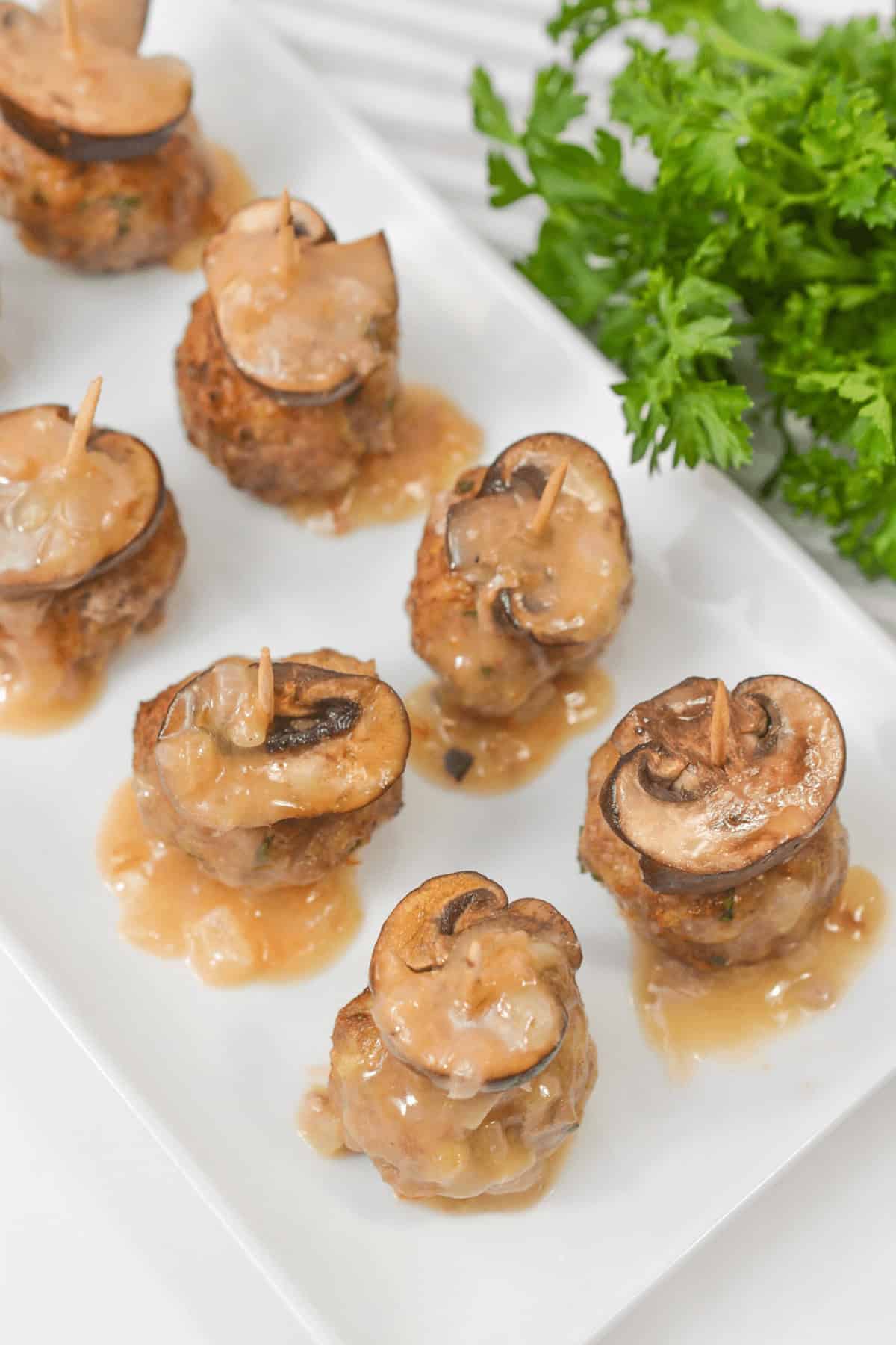 Savoury food with mushroom slices and skewered with toothpicks, drizzled with sauce, are arranged on a rectangular white plate. Fresh parsley is in the background.