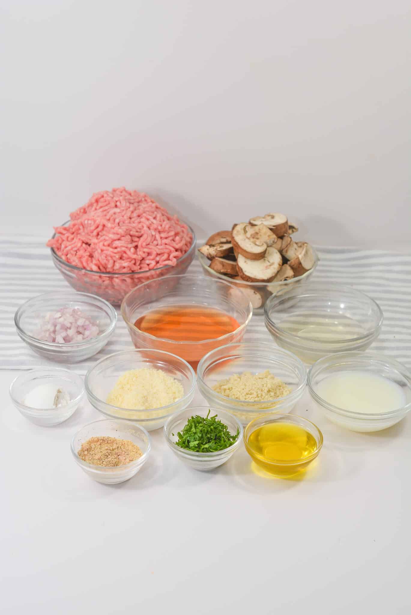 A delightful array of ingredients in clear bowls, featuring ground meat, sliced mushrooms, chopped onions, grated cheese, breadcrumbs, herbs, oil, and liquids—all beautifully arranged on a pristine white surface.