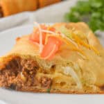 A close-up of a Taco Crescent Ring slice on a white plate, topped with shredded cheese, tomatoes, and lettuce, set against a blurred background.
