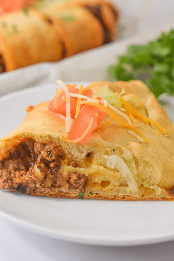 A close-up of a Taco Crescent Ring slice on a white plate, topped with shredded cheese, tomatoes, and lettuce, set against a blurred background.