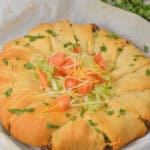 A Taco Crescent Ring brimming with beef and topped with shredded lettuce, diced tomatoes, and cheese rests on parchment paper, garnished with fresh cilantro.