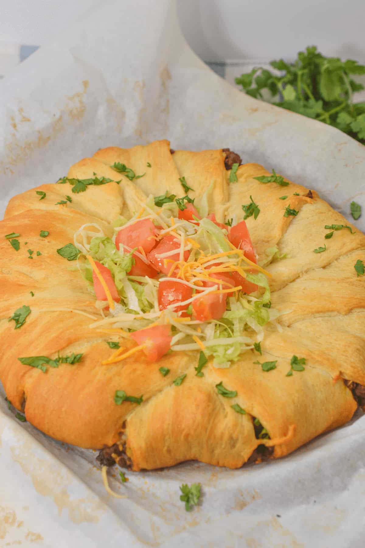 A Taco Crescent Ring brimming with beef and topped with shredded lettuce, diced tomatoes, and cheese rests on parchment paper, garnished with fresh cilantro.