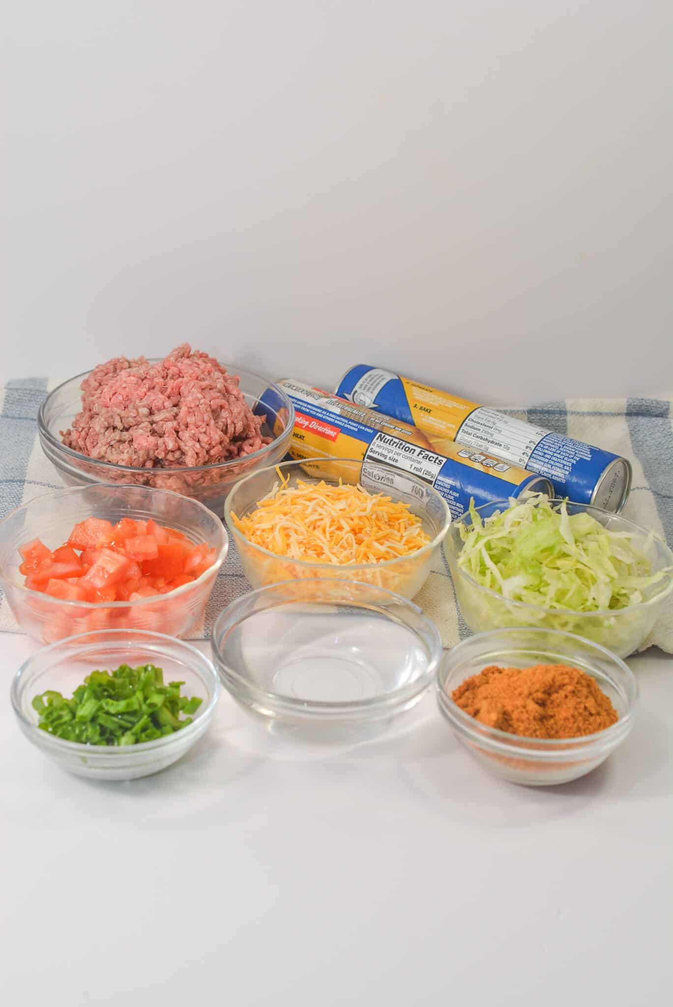 Ingredients for the Taco Crescent Ring include ground beef, chopped tomatoes, shredded cheese, shredded lettuce, green onions, taco seasoning, crescent roll dough, and a small bowl of water on a cloth.
