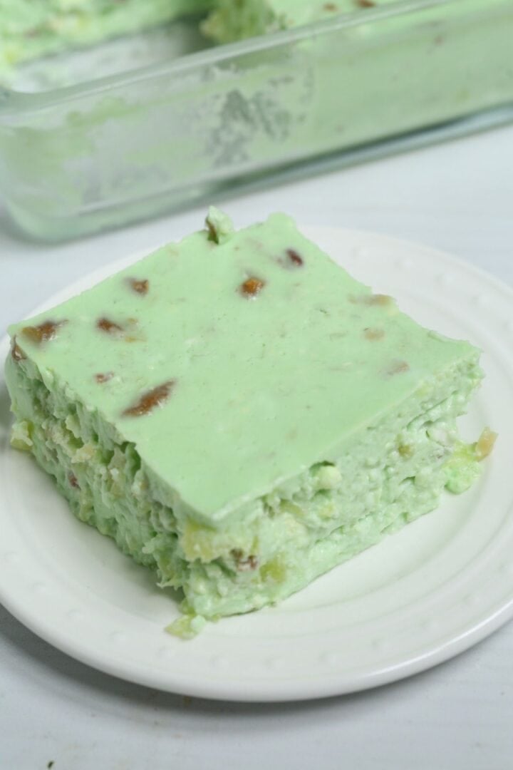 A square slice of pale green cottage cheese jello salad with fruit pieces, served on a white plate.