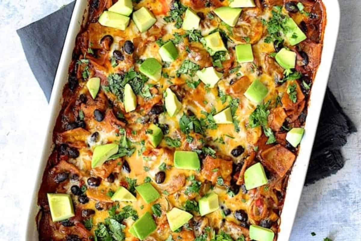 A baked casserole inspired by seasonal recipes, topped with melted cheese, black beans, chopped avocado, and fresh cilantro, served in a white rectangular dish.