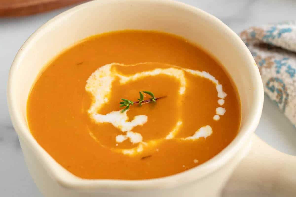 A bowl of orange-colored squash soup, garnished with a swirl of cream and a small sprig of fresh thyme on top—a perfect choice for lovers of seasonal recipes.