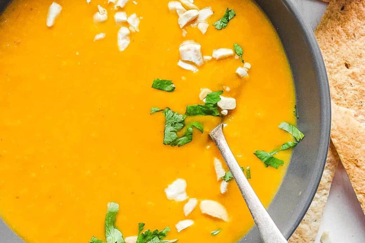 A bowl of orange soup, perfect for squash recipes, garnished with chopped cashews and fresh cilantro, with a spoon inside and a piece of bread on the side.