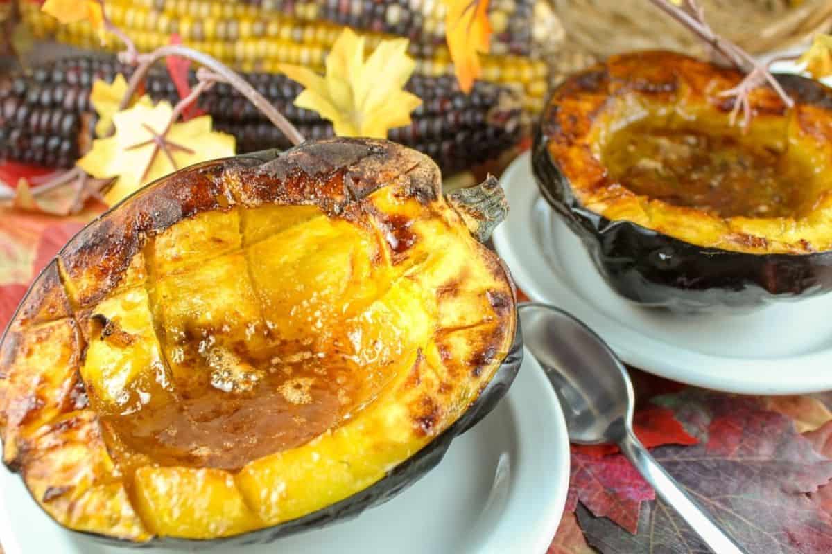 Two halves of baked acorn squash with a brown sugar glaze are served on white plates, spoons beside each, with autumn leaves and corn in the background—a perfect choice for fans of seasonal recipes and comforting squash dishes.