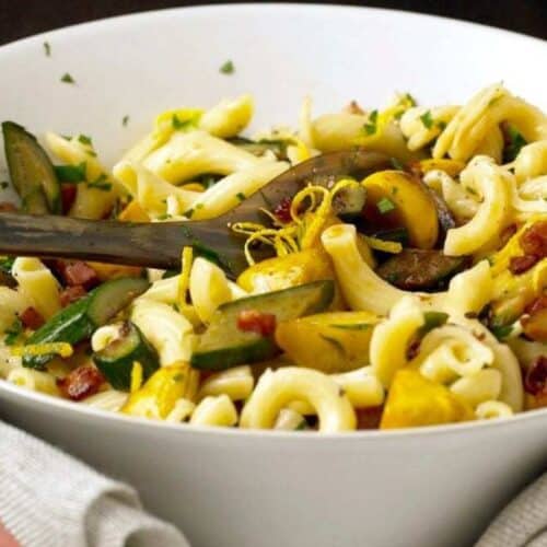 A bowl of macaroni pasta mixed with zucchini, yellow squash, sun-dried tomatoes, herbs, and lemon zest—perfect for those who love fresh squash recipes—with a serving spoon inside.