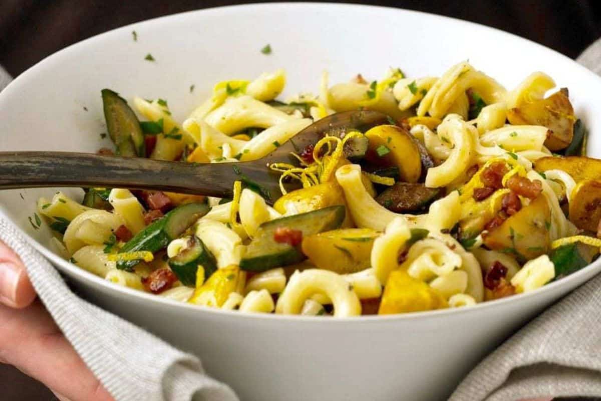 A bowl of macaroni pasta mixed with zucchini, yellow squash, sun-dried tomatoes, herbs, and lemon zest—perfect for those who love fresh squash recipes—with a serving spoon inside.