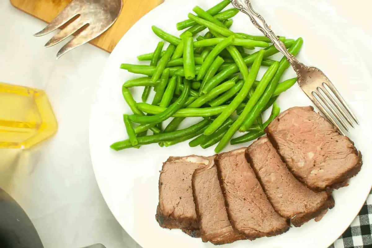 A white plate with five slices of air fryer roast beef and a serving of green beans, with a fork resting on the plate—a classic choice from Rib Roast Recipes sure to impress guests at any special gathering.