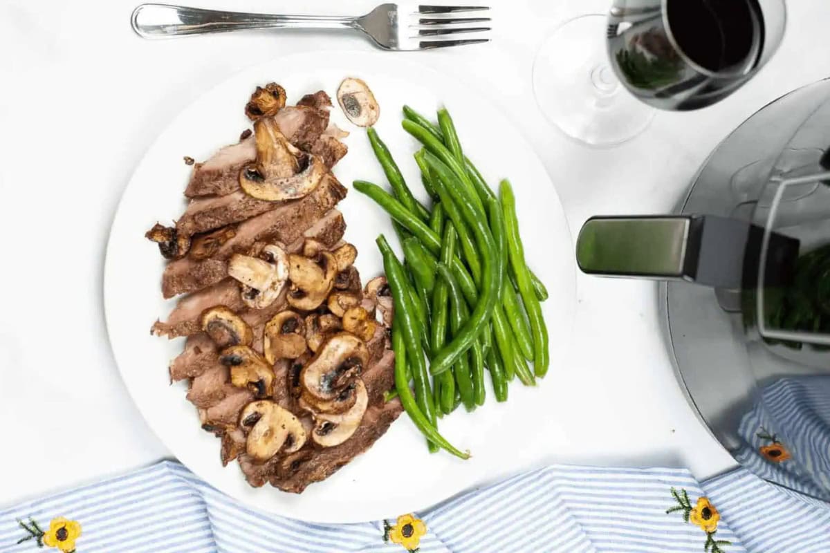 A white plate with sliced ribeye steak topped with mushrooms and served with green beans, sits next to a fork and a glass of dark red wine—perfect to impress guests at your next gathering.