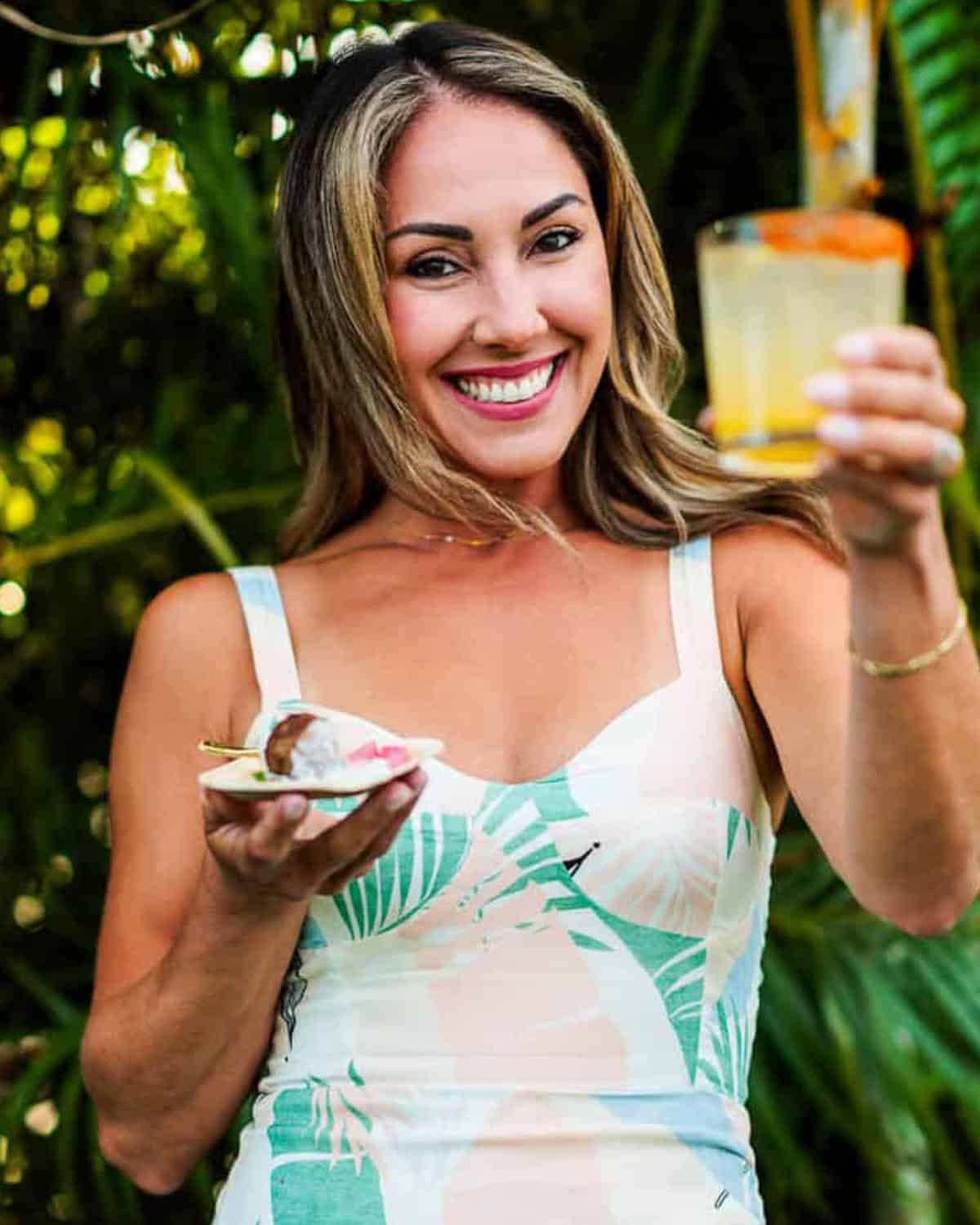 A smiling woman effortlessly entertains outdoors, holding a drink in her right hand and a small plate of food in her left, surrounded by lush greenery that adds a touch of calm and less stress to the gathering.