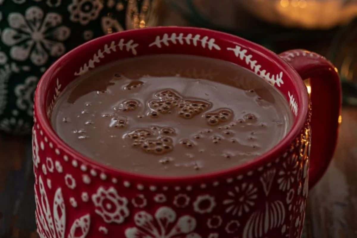 A red mug with white floral patterns filled with rich hot cocoa sits on a wooden surface, steam rising from the frothy beverage.