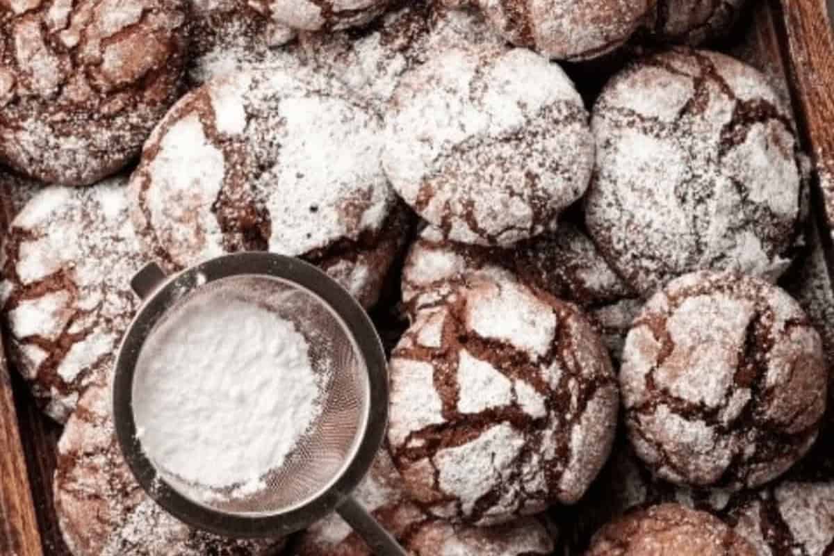 A tray of chocolate brownie crinkle cookies dusted with powdered sugar with a metal sieve containing powdered sugar resting on top.