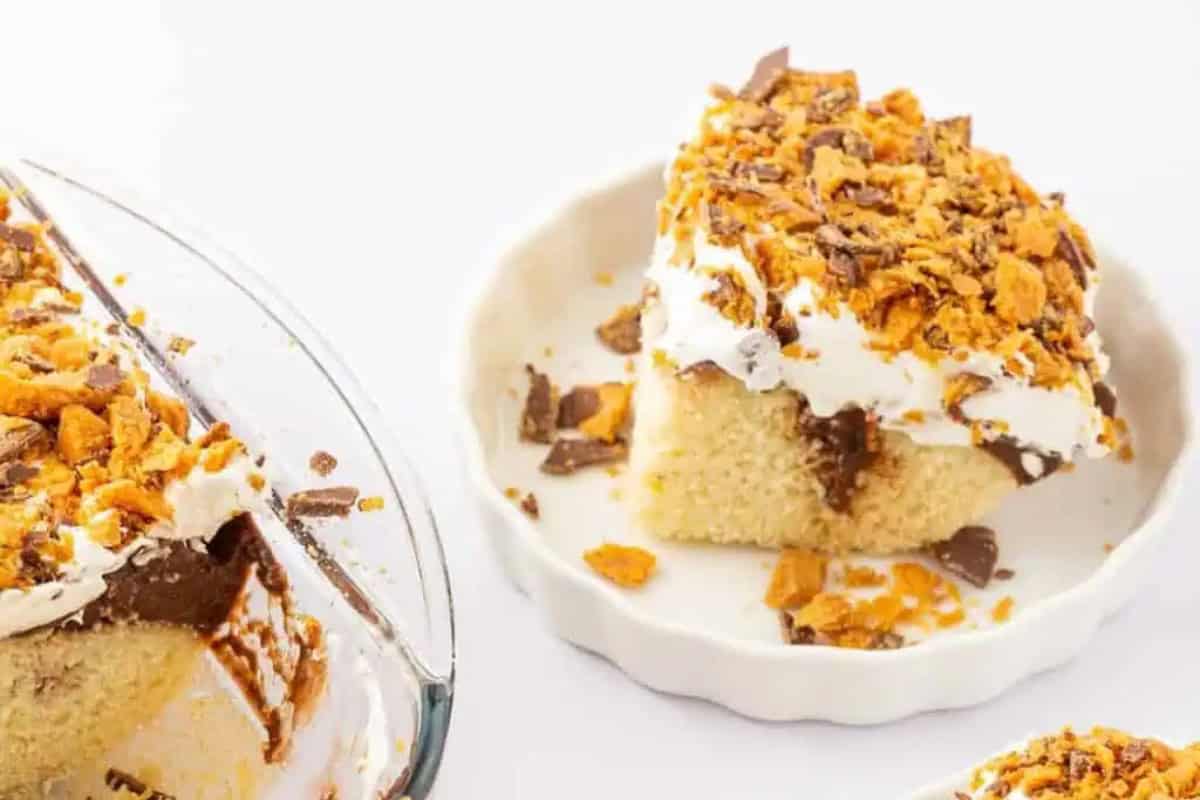 A slice of yellow cake topped with whipped cream, chocolate, and crushed butterfingers sits on a white plate next to a pan of the rest of the cake.