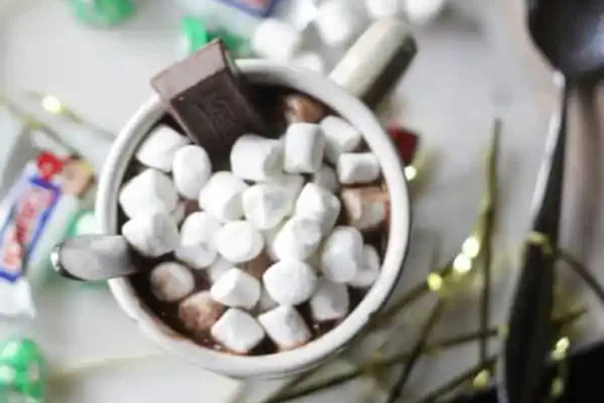 A mug of creamy hot cocoa topped with mini marshmallows and a piece of chocolate, surrounded by candies and a spoon—perfect for warm drinks or trying new hot cocoa recipes.