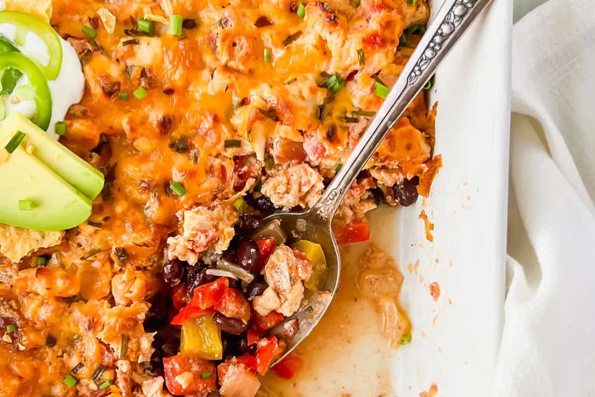 A close-up of a comforting chicken casserole with melted cheese, beans, vegetables, and a metal serving spoon. Sliced avocado and jalapeño are garnished on top, making this dish the ultimate comfort food.