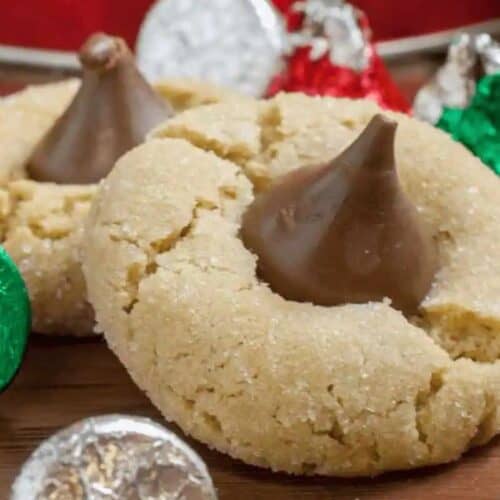 Two cozy cookies, each topped with a chocolate candy, are surrounded by foil-wrapped chocolates on a wooden surface—perfect Christmas cookies for sharing or adding to your make ahead cookie recipes collection.