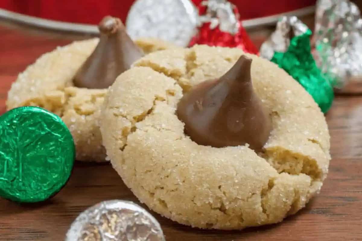 Two peanut butter cookies, each topped with a chocolate candy kiss, are surrounded by foil-wrapped chocolate kisses on a wooden surface