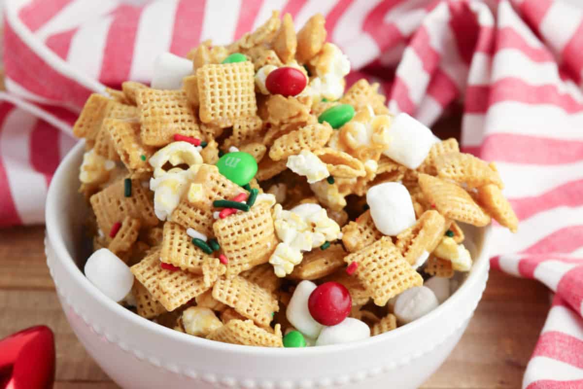A white bowl filled with Chex mix that consists of Chex, mini marshmallows, popcorn, red and green chocolate candies, and sprinkles set against a festive red and white striped cloth background..