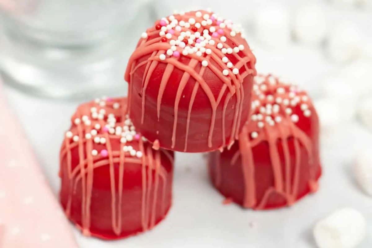 Round chocolate hot cocoa bombs with marshmallows on the side, displayed on a white surface.