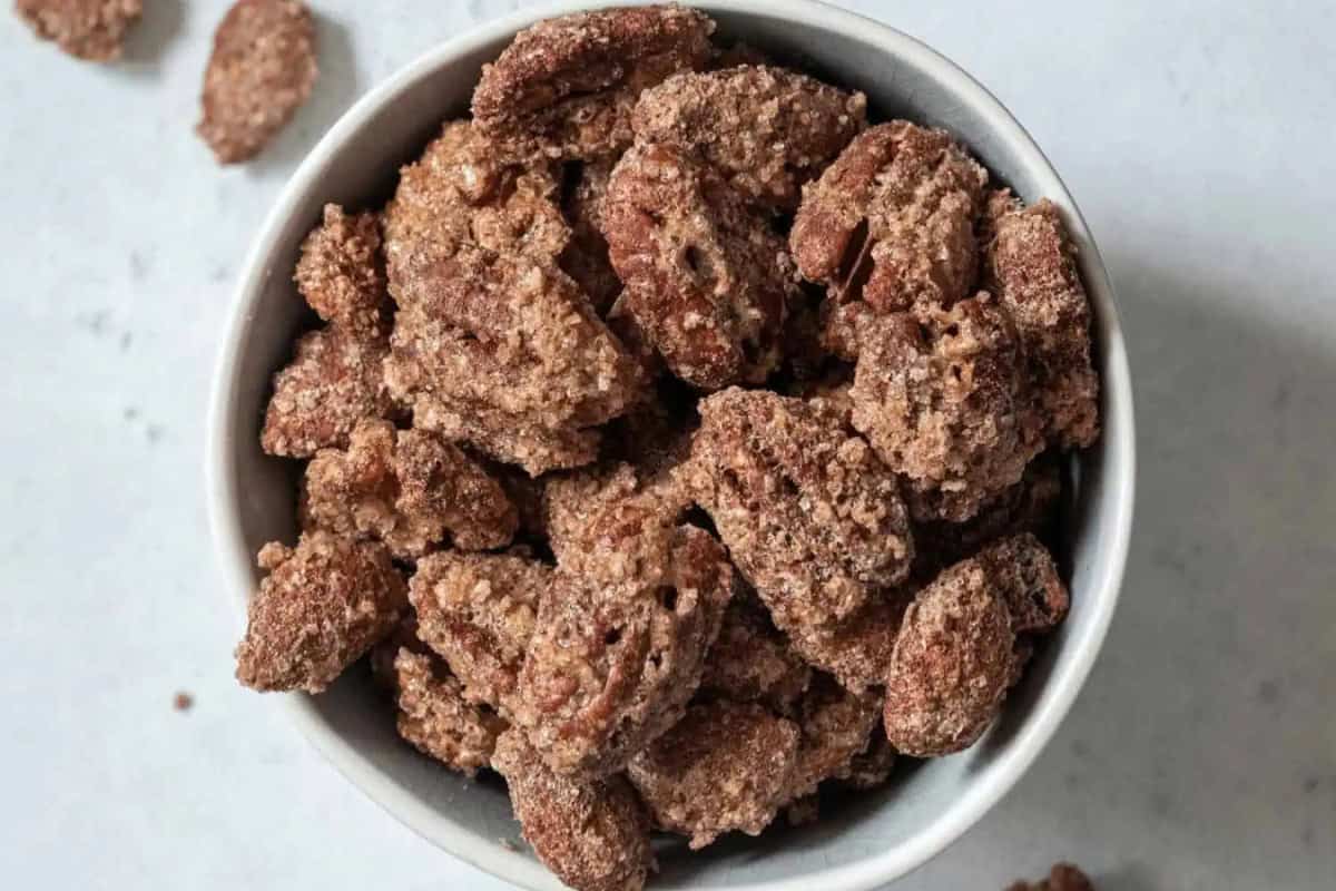 A white bowl filled with sugared roasted cinnamon pecans sits on a light surface with a few nuts scattered nearby.