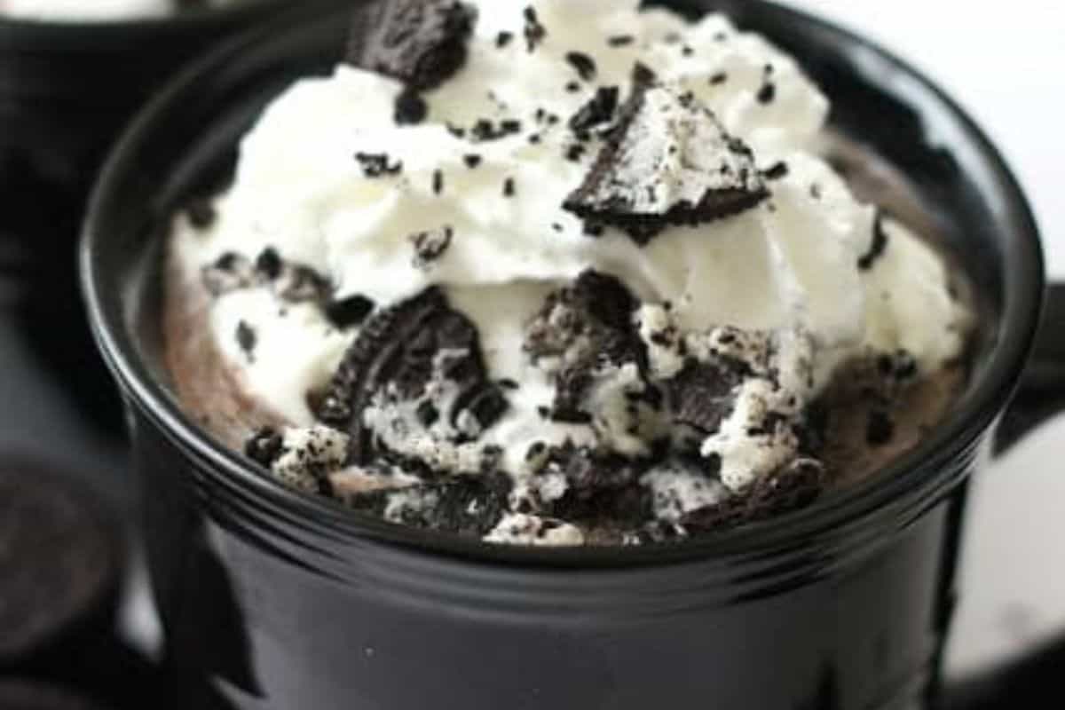 A black mug filled with rich hot chocolate, topped with whipped cream and crumbled chocolate sandwich cookies.