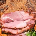 Close-up of a copycat honey baked ham with several slices cut, showcasing its juicy pink interior. Grilled orange halves and greens are visible in the foreground.