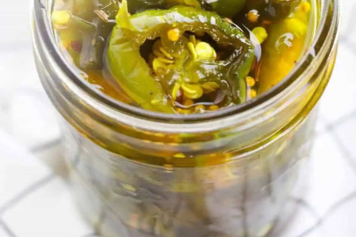 A glass jar filled with sliced pickled candied jalapeño peppers in brine, viewed from above on a white surface.