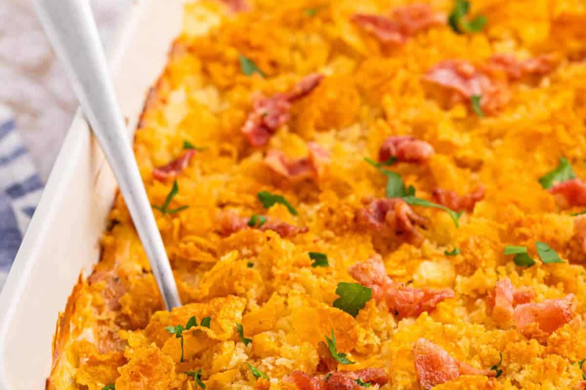 Close-up of a baked comfort food casserole, topped with crispy crushed cornflakes, bacon pieces, and parsley in a white baking dish with a serving spoon—a delicious twist on classic chicken casseroles.