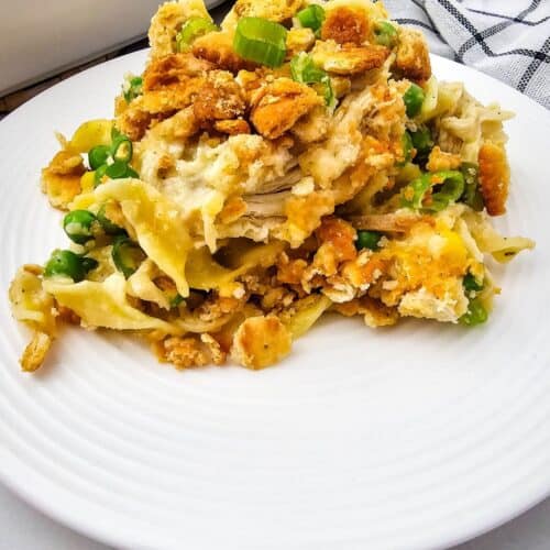 A serving of Creamy Chicken Noodle Casserole with noodles, shredded chicken, green peas, and a golden crumb topping sits on a white plate, with a striped cloth in the background.
