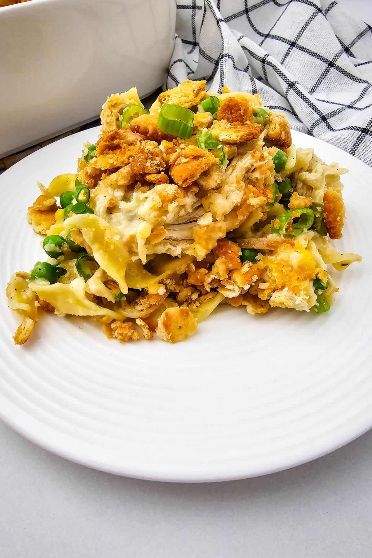 A serving of Creamy Chicken Noodle Casserole with noodles, shredded chicken, green peas, and a golden crumb topping sits on a white plate, with a striped cloth in the background.