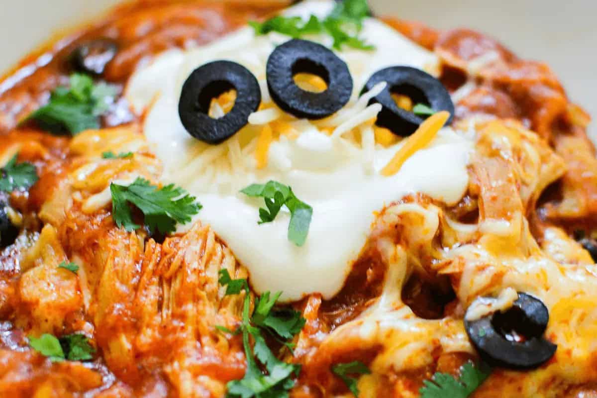 Close-up of a cheesy chicken enchilada, classic comfort food, topped with sour cream, sliced black olives, shredded cheese, and chopped cilantro—a must-try for fans of chicken casseroles.