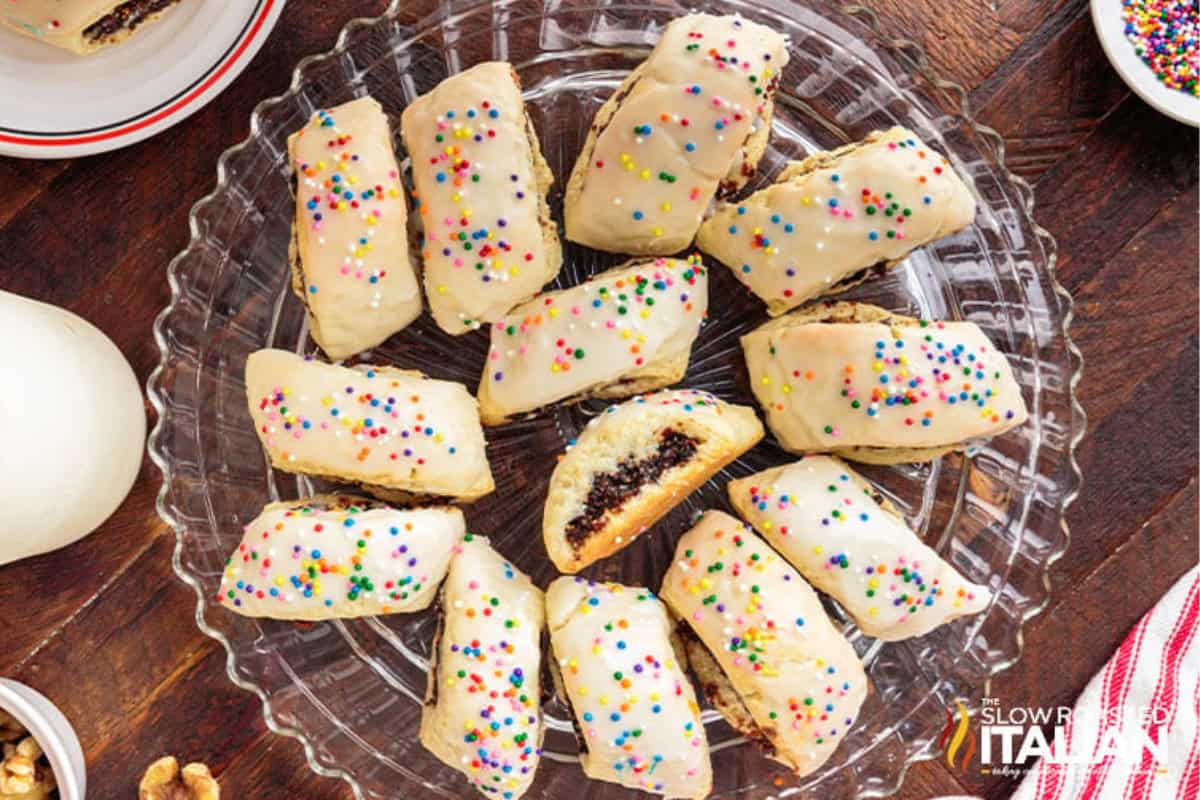 A glass platter with twelve rectangular Cuccidati holiday cookies with colorful sprinkles and glazed with icing arranged in a circular pattern on a wooden table. One cookie is cut open to reveal a rich dark fig filling.