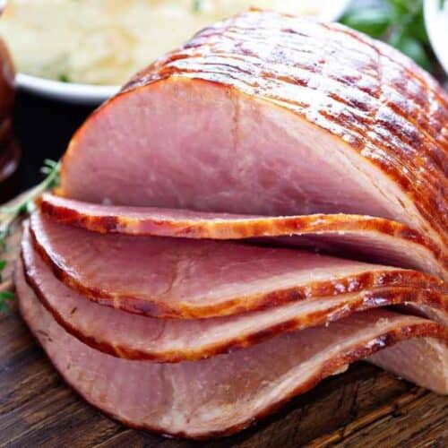 Sliced glazed ham on a wooden cutting board with herbs, pepper mills, and side dishes in the background—a perfect centerpiece for a family feast or inspiration for your next ham recipes.
