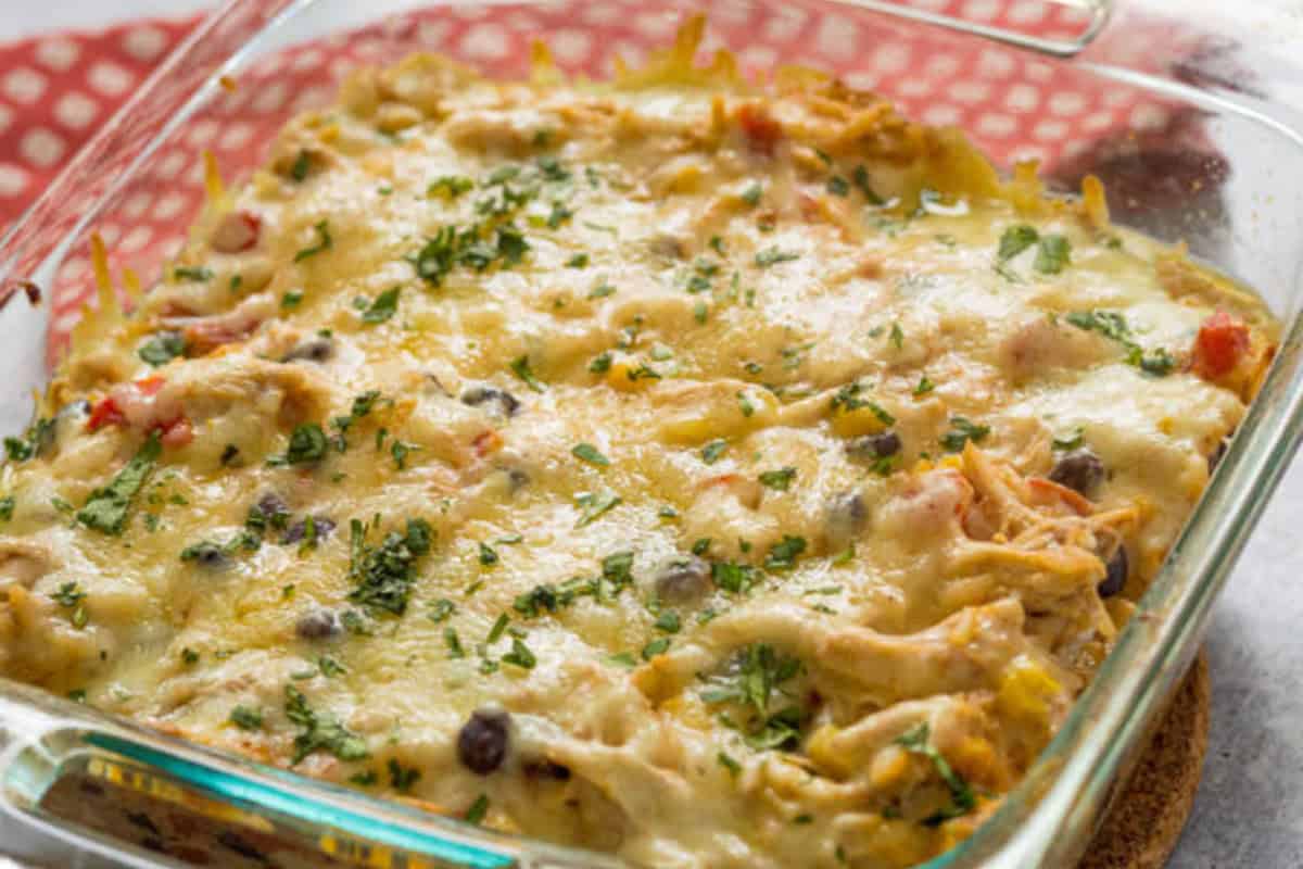 A glass baking dish filled with a comfort food classic: a baked chicken casserole topped with melted cheese, black beans, and chopped herbs, resting on a red patterned cloth.