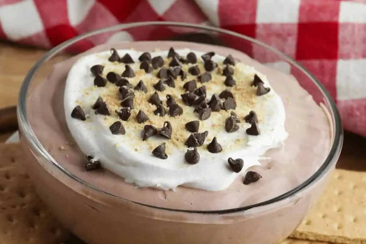 A glass bowl filled with chocolate mousse, topped with whipped cream, chocolate chips, and graham cracker crumbs, sits on a wooden surface with a red plaid cloth and graham crackers/