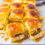 Breakfast sliders featuring scrambled eggs, sausage, and melted cheese on a baking sheet, all perfectly drizzled with syrup.
