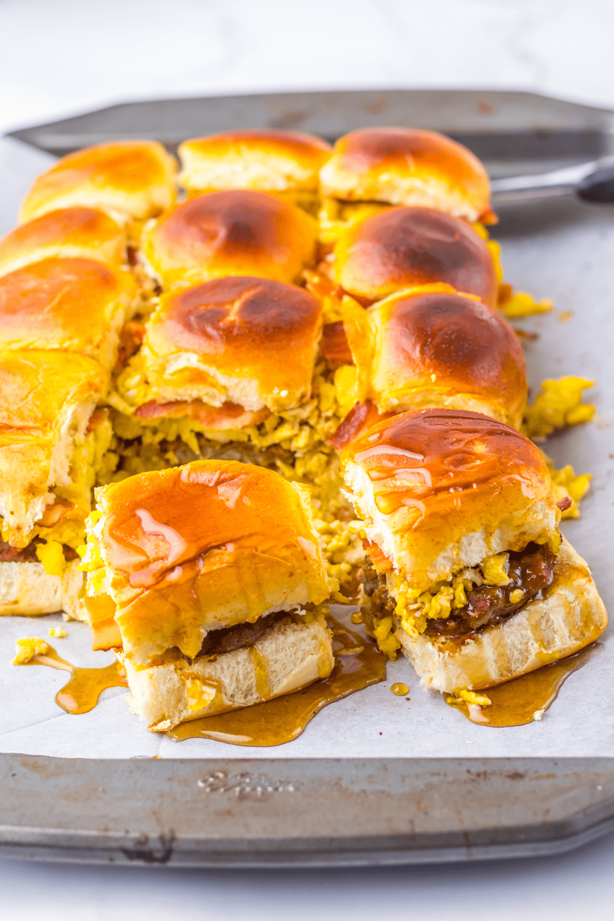 Breakfast sliders featuring scrambled eggs, sausage, and melted cheese on a baking sheet, all perfectly drizzled with syrup.