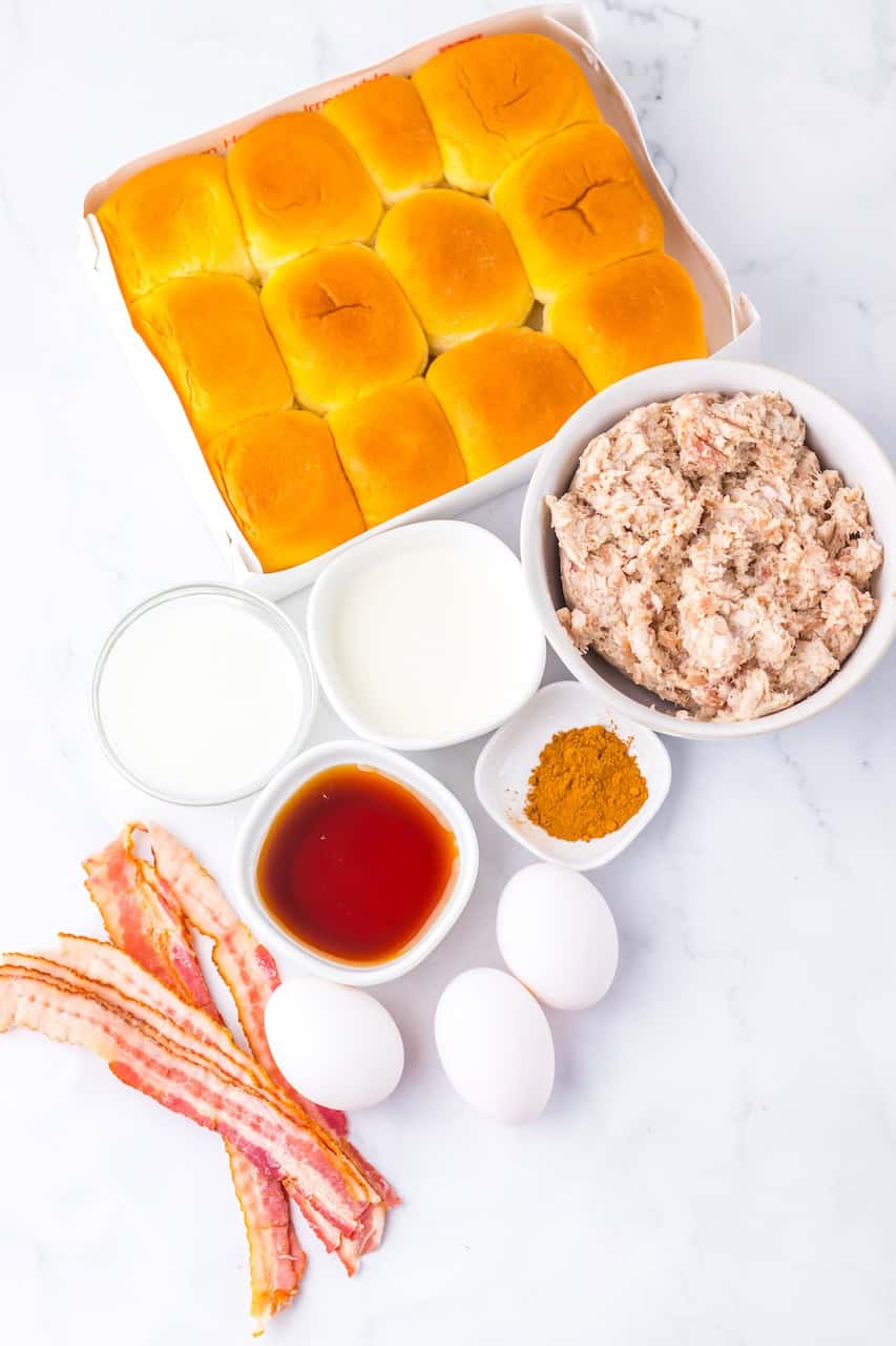 Ingredients for delicious breakfast on a white surface: tray of rolls, bowl of minced meat, bacon strips, three eggs, bowls of milk, soy sauce, and curry powder.