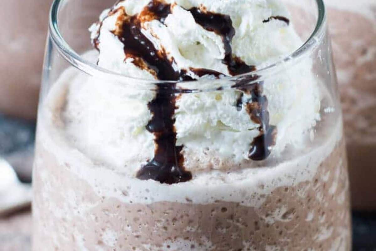 A glass of blended frozen hot chocolate topped with whipped cream and a drizzle of chocolate syrup, inspired by the decadence of creamy hot cocoa.