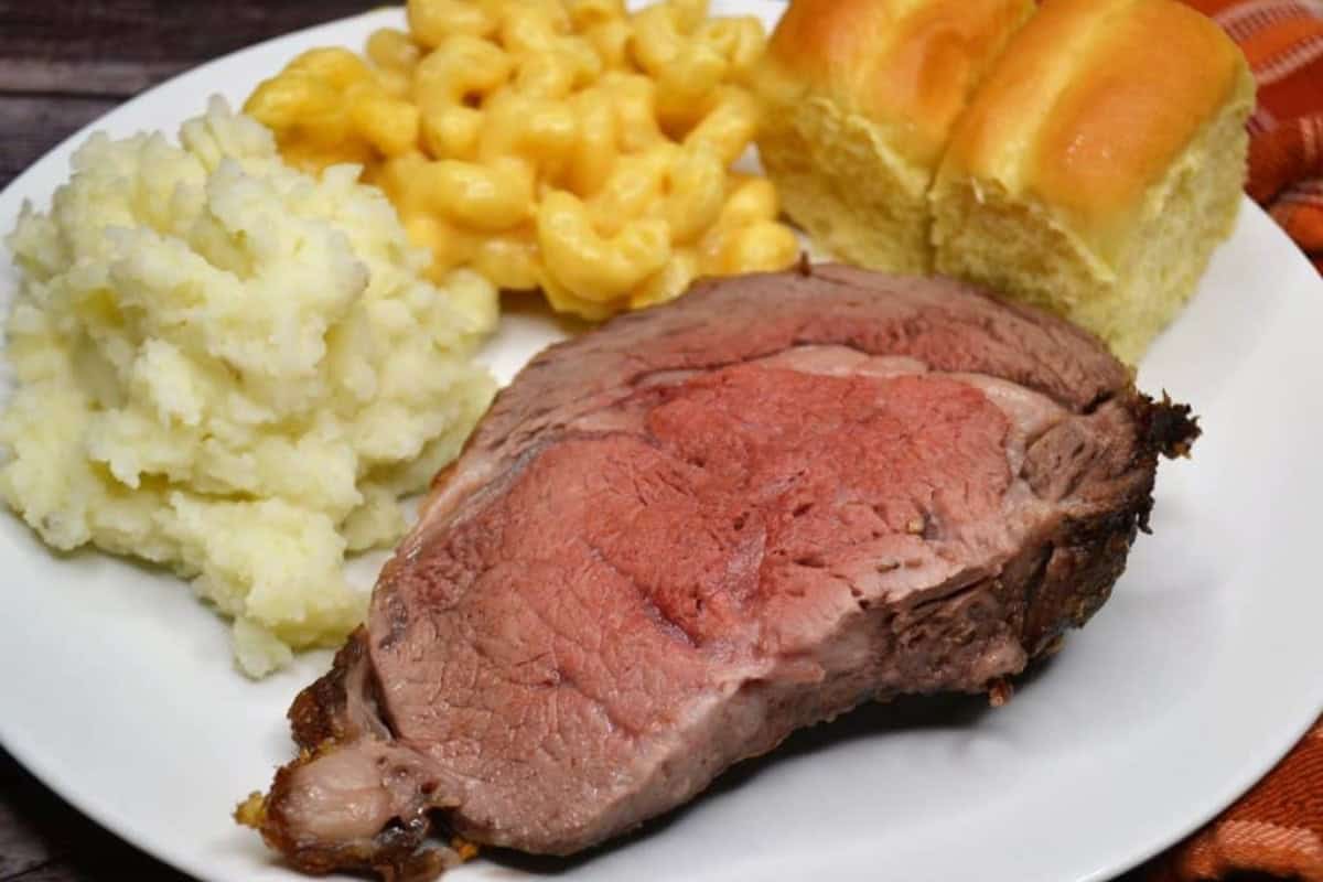 A plate with a slice of grilled prime rib roast beef f, mashed potatoes, macaroni and cheese, and two dinner rolls.