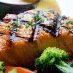 Grilled Salmon fillet with crosshatch grill marks, garnished with herbs, served with steamed broccoli, sliced carrots, lemon wedges, and a small cup of sauce in the background—a delightful Grilled Salmon dish that’s both vibrant and fresh.